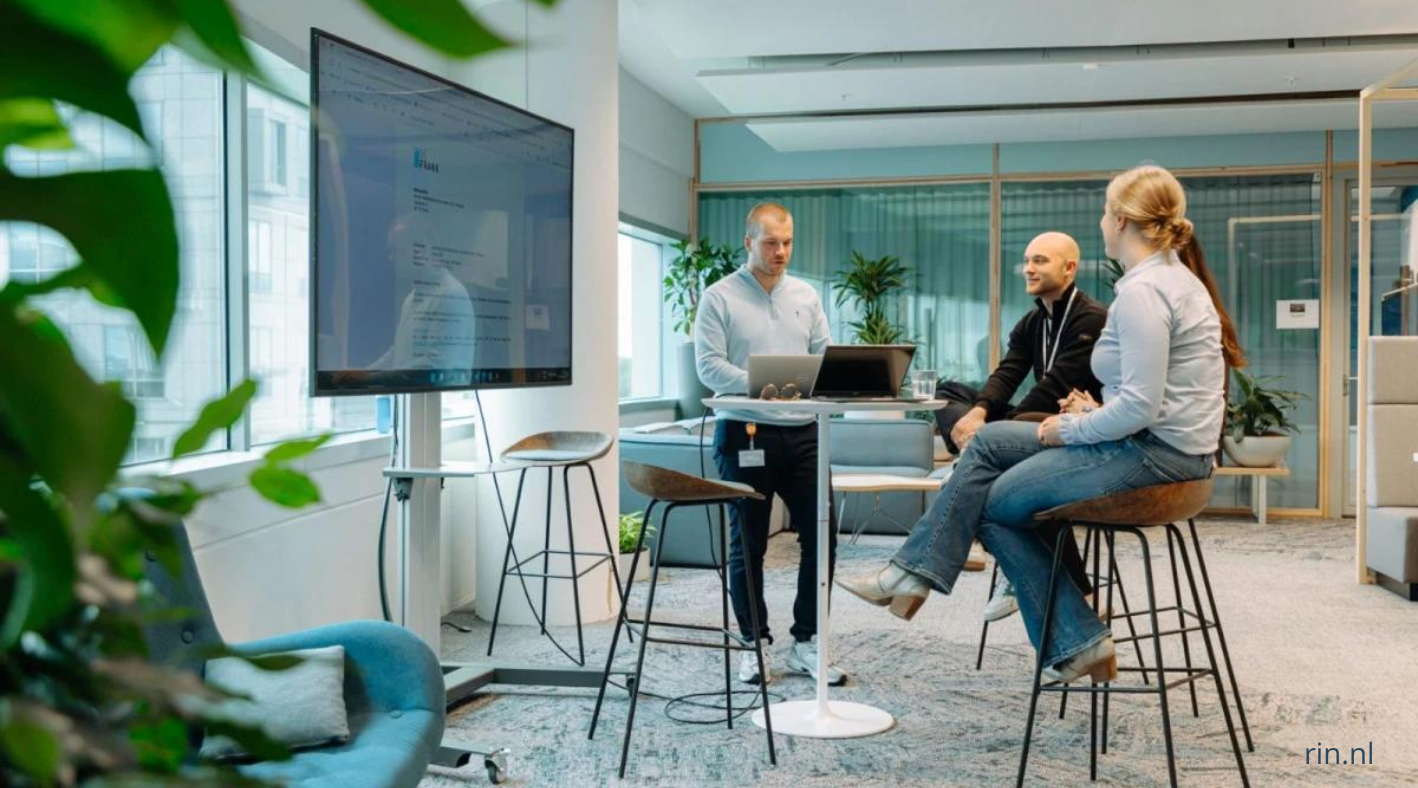 Medewerkers aan het werk in een moderne kantooromgeving, met laptops en beeldschermen op verschillende werkplekken en overleg tussen collega’s.